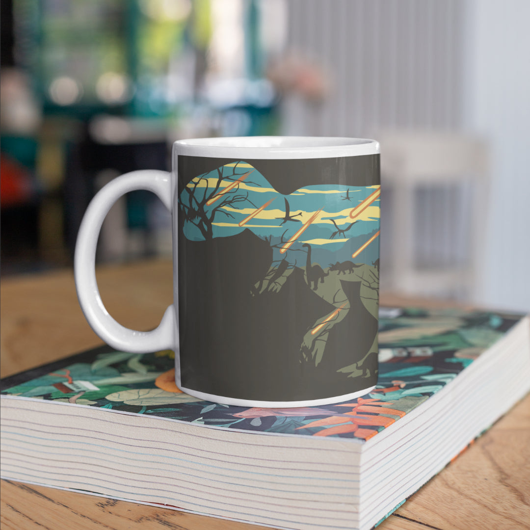 A dinosaur mug sitting on a book about dinosaurs. The mug features a dark grey background, with a stencil of a T-Rex. Inside the stencil of the T-Rex shows the final event of the dinosaurs with meteors raining down. 
